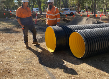 Two construction workers in high-visibility clothing standing beside large Iplex BlackMax corrugated drainage pipes at a Keystone Civil construction site